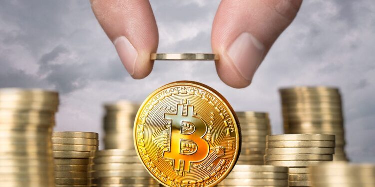 monedas físicas de bitcoin doradas apiladas una encima de otra, mientras dos dedos sostienen otra moneda de bitcoin.