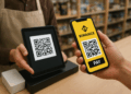 Una persona se encuentra en una tienda realizando un pago con su teléfono móvil. En la pantalla del celular se ve claramente el logotipo amarillo de Binance y un código QR con el botón “PAY” debajo. Frente a ella, un empleado sostiene una tableta negra que muestra otro código QR para recibir el pago. Ambos están en un mostrador de madera, con estantes llenos de productos desenfocados al fondo. La iluminación es cálida y natural, dando un ambiente acogedor y cotidiano, típico de una tienda moderna.