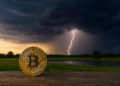 Moneda de Bitcoin en primer plano sobre una superficie de madera, brillando con tonos dorados. El entorno se muestra tranquilo, con un campo verde y un lago detrás. Al fondo, una tormenta comienza a formarse con nubes oscuras y un rayo iluminando el cielo.