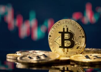 Bitcoin sobre una mesa y gráficas de mercado en el fondo.