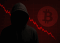 Una escena oscura y realista. En primer plano hay una persona con una sudadera negra con capucha, de pie, con la cara completamente en sombra, sin rasgos visibles. Transmite una sensación de anonimato y misterio, representando simbólicamente a Satoshi Nakamoto, el creador anónimo de Bitcoin. En el fondo, se ve un gráfico financiero en color rojo que desciende bruscamente, indicando una fuerte caída del precio de Bitcoin. Sobre el gráfico, se distingue el logotipo de Bitcoin en tono rojizo y difuso, como si estuviera iluminado detrás de la figura. Todo el ambiente está teñido de tonos oscuros y rojos, evocando tensión y un sentimiento de pérdida o declive.