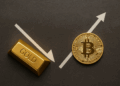 Imagen horizontal que muestra una barra de oro y una moneda de bitcoin sobre un fondo oscuro. Una flecha descendente apunta hacia el lingote de oro, simbolizando la caída de su precio, mientras otra flecha ascendente se dirige hacia el bitcoin, representando su aumento de valor.