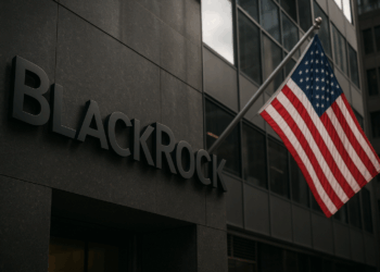 Imagen realista de la sede de BlackRock en Estados Unidos. En primer plano se observa el logotipo metálico de la compañía sobre la fachada de un edificio moderno, con una bandera estadounidense ondeando a la derecha. El entorno urbano refleja un clima financiero y corporativo.