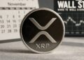 Una moneda física de XRP en primer plano, metálica con el logo blanco estilizado y la inscripción "XRP" grabada, reposa sobre una superficie de madera. En el fondo ligeramente desenfocado, un calendario de escritorio muestra el mes de noviembre y, a su lado, la señalización negra de "Wall Street".