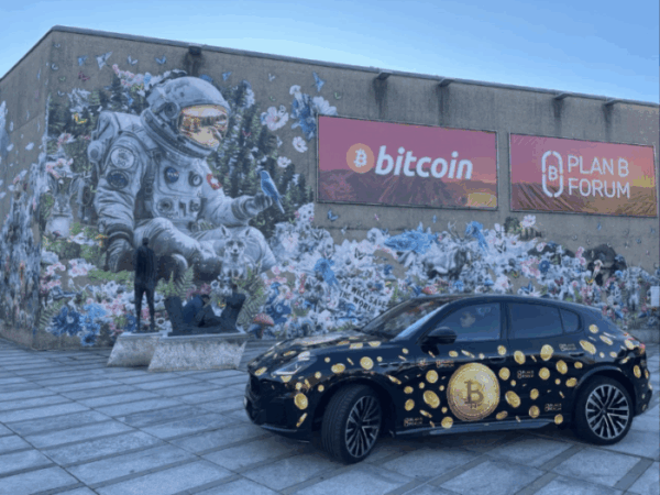 Vehículo con el logo de bitcoin. de fondo un mural con vallas alusivas a evento bitcoiner Plan B.