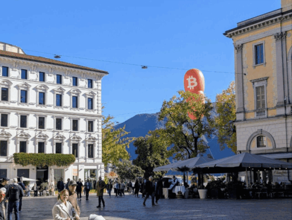 Plaza con edificios alrededor y al fondo un globo gigante color naranja con logo de bitcoin en Lugano. Suiza.