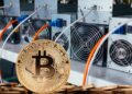 Moneda física de bitcoin dorada con mineros ASIC detrás.