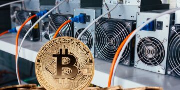 Moneda física de bitcoin dorada con mineros ASIC detrás.