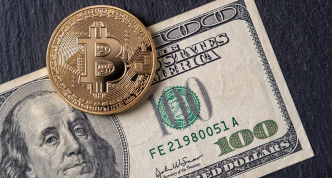 Moneda física de bitcoin dorada sobre un billete de 100 dólares sobre una mesa.