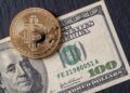 Moneda física de bitcoin dorada sobre un billete de 100 dólares sobre una mesa.