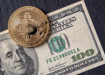 Moneda física de bitcoin dorada sobre un billete de 100 dólares sobre una mesa.