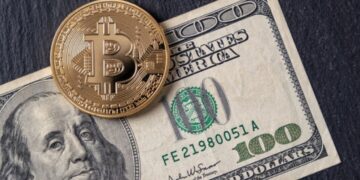 Moneda física de bitcoin dorada sobre un billete de 100 dólares sobre una mesa.