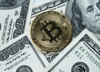 Moneda física de bitcoin dorada rodeada de billetes físicos de 100 dólares.