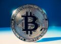 Moneda física de bitcoin dorada sobre una mesa blanca y fondo azul.