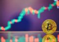 Moneda física de bitcoin doarada sobre un espejo y con gráficas de mercado en el fondo.