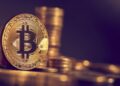 Moneda física dorada de bitcoin con otras monedas apiladas detrás en fondo púrpura.