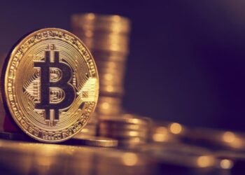 Moneda física dorada de bitcoin con otras monedas apiladas detrás en fondo púrpura.