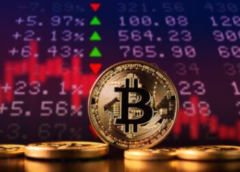 Monedas físicas de bitcoin doradas sobre un escritorio con una gráfica en rojo y números en el fondo.