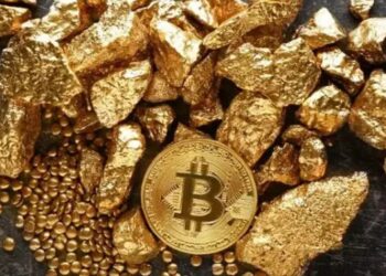 Una moneda física de bitcoin dorada rodeada de trozos de oro sobre una mesa.