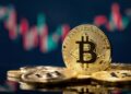 Monedas físicas de bitcoin en un escritorio y un fondo negro con gráficas de mercado verdes y rojas.