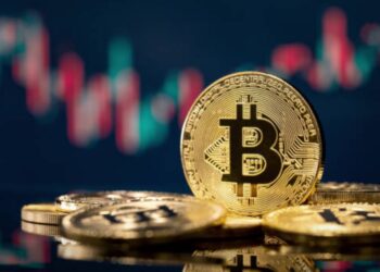 Monedas físicas de bitcoin en un escritorio y un fondo negro con gráficas de mercado verdes y rojas.