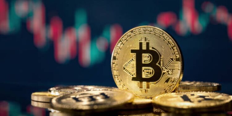 Monedas físicas de bitcoin en un escritorio y un fondo negro con gráficas de mercado verdes y rojas.