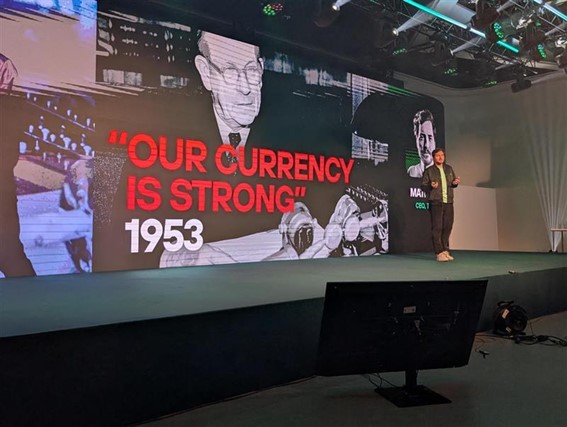 A man gives a presentation on an illuminated stage. Behind him is a large screen with a black and white image and the phrase in red “Our currency is strong.”