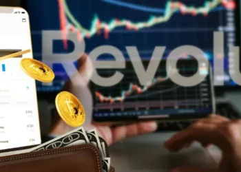 Logo de Revolut sobre un smartphone y una laptop con indicadores de mercados.