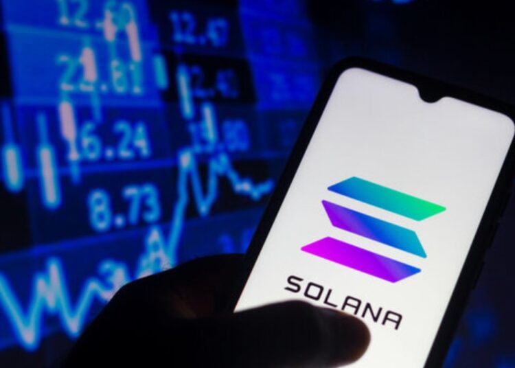 Una mano sujetando un teléfono inteligente que muestra el logo de Solana, y de fondo una pared de gráficas en color azul.