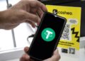 Un teléfono inteligente sostenido por unas manos, el teléfono muestra el logo de USDT y en el fondo hay un cartel de Cashea.