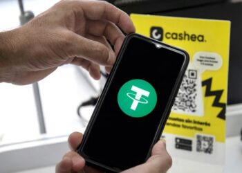 Un teléfono inteligente sostenido por unas manos, el teléfono muestra el logo de USDT y en el fondo hay un cartel de Cashea.