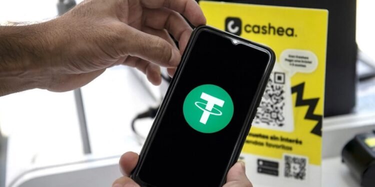 Un teléfono inteligente sostenido por unas manos, el teléfono muestra el logo de USDT y en el fondo hay un cartel de Cashea.