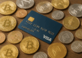 Una tarjeta de crédito Visa azul sobre una mesa de madera, rodeada de varias criptomonedas físicas en forma de monedas doradas, plateadas y cobrizas, entre ellas bitcoin, ethereum y litecoin.