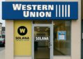 Vista frontal realista y horizontal de una sucursal de Western Union en una calle urbana. El letrero principal muestra "Western Union" en azul con letras blancas. En la ventana izquierda aparece el logo de Solana integrado con el de Western Union, incluyendo el texto "W SOLANA THE NAIOR NORIBE". En la puerta de vidrio central se ve el logo de "SOLANA" en letras negras. Al fondo, se observa el interior iluminado de la oficina, con un auto pasando en la calle.