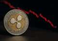 Una moneda física de Ripple (XRP) en primer plano, con diseño dorado y plateado, y al fondo un gráfico de velas rojas en caída que representa la bajada de su precio en el mercado.