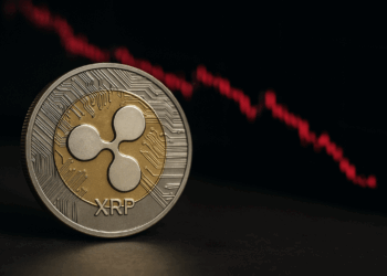 Una moneda física de Ripple (XRP) en primer plano, con diseño dorado y plateado, y al fondo un gráfico de velas rojas en caída que representa la bajada de su precio en el mercado.