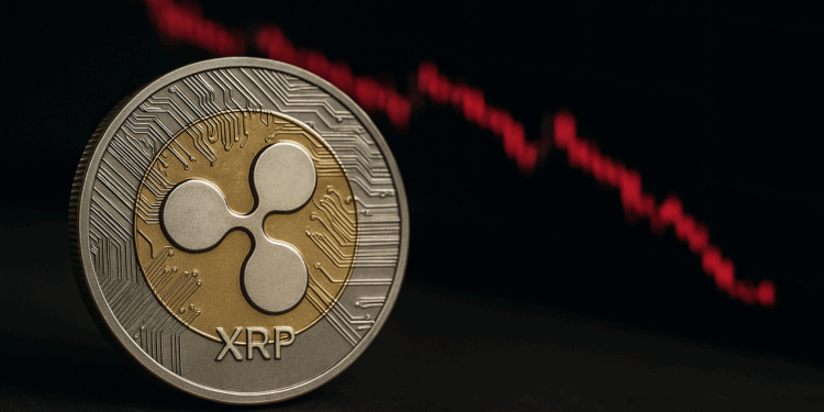 Una moneda física de Ripple (XRP) en primer plano, con diseño dorado y plateado, y al fondo un gráfico de velas rojas en caída que representa la bajada de su precio en el mercado.