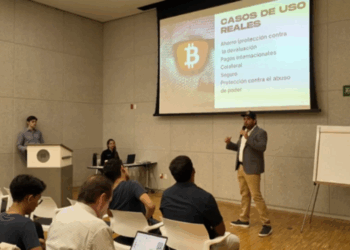 Un grupo de personas reciben charla sobre bitcoin en un salón con una pantalla en el que se observa el logo de BTC.