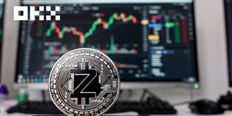 una moneda plateada de zcash, junto a una interfaz de trading en la pantalla de un computadora y el logo del exchange OKX.