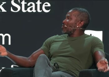 Arthur Hayes vistiendo una camiseta verde y pantalón gris, mientras habla en un escenario durante una conferencia.
