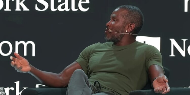 Arthur Hayes vistiendo una camiseta verde y pantalón gris, mientras habla en un escenario durante una conferencia.