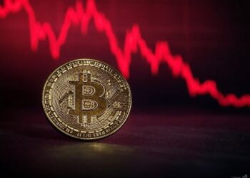 Una moneda física de bitcoin delante de un gráfico de precios en rojo.
