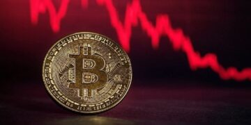 Una moneda física de bitcoin delante de un gráfico de precios en rojo.