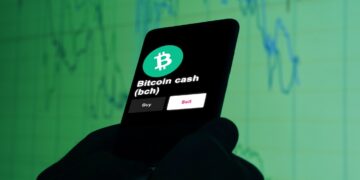 Un celular con el logo de la cripmoneda bitcoin cash delante de gráficos de precios.