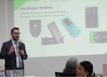 Una persona usa micrófono para dirigirse a un público. A su espalda se observa una imagen proyectada de wallets frías de bitcoin.