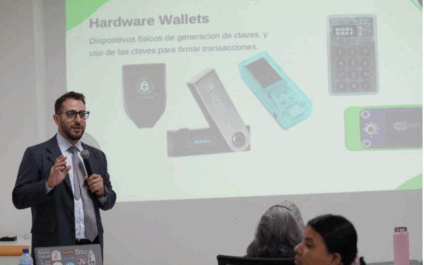 Una persona usa micrófono para dirigirse a un público. A su espalda se observa una imagen proyectada de wallets frías de bitcoin.