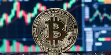 Una moneda física de bitcoin con un fondo con gráficos bursátiles en rojo y verde.