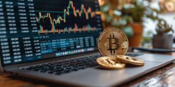 Moneda física de bitcoin dorada sobre una laptop que está mostrando gráficas de mercado y está sobre un escritorio.
