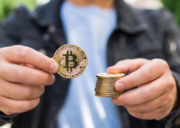 Hombre sostiene una moneda física de bitcoin en una mano y una pila de monedas también de bitcoin en la otra.