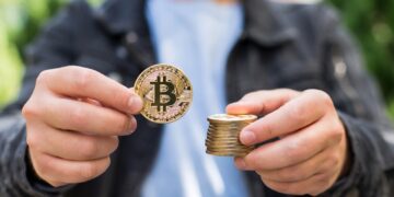 Hombre sostiene una moneda física de bitcoin en una mano y una pila de monedas también de bitcoin en la otra.
